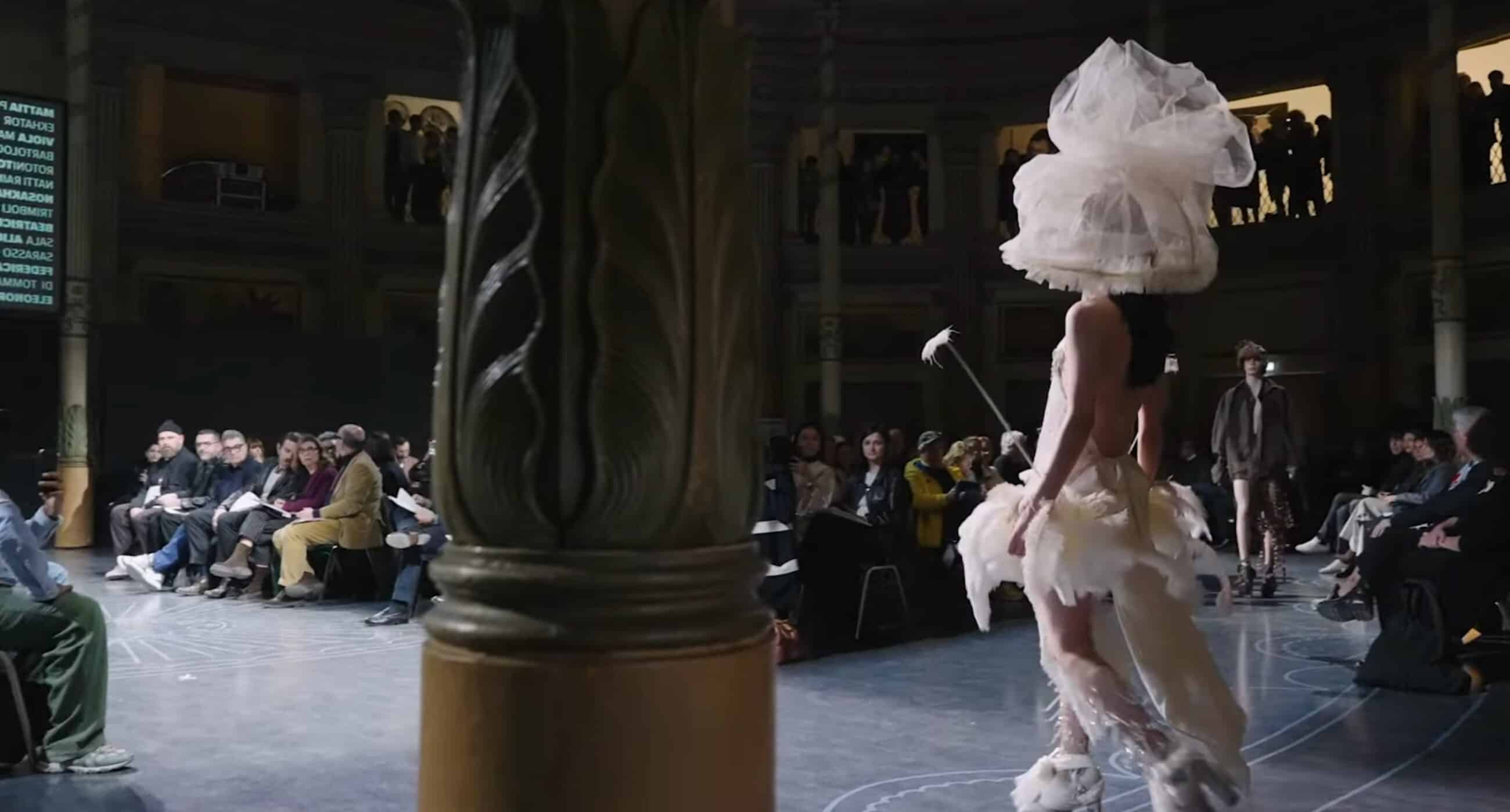 A model walks down a circular runway wearing a dramatic white outfit with a large headpiece. Seated spectators surround the runway, with a decorative column in the foreground. This appears to be a fashion show in a grand, multi-story venue showcasing trends seen in top online fashion courses.