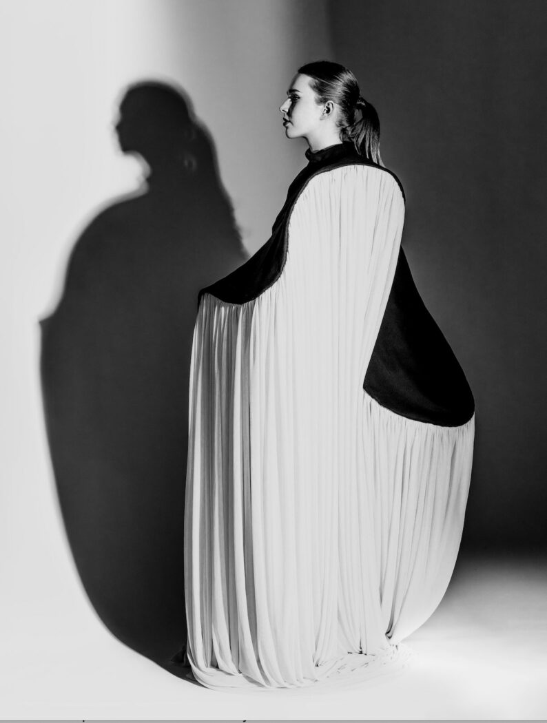 A woman stands in profile against a plain backdrop, wearing a long, flowing monochrome dress that casts a prominent shadow on the wall, as if preparing for the next chapter in her career.