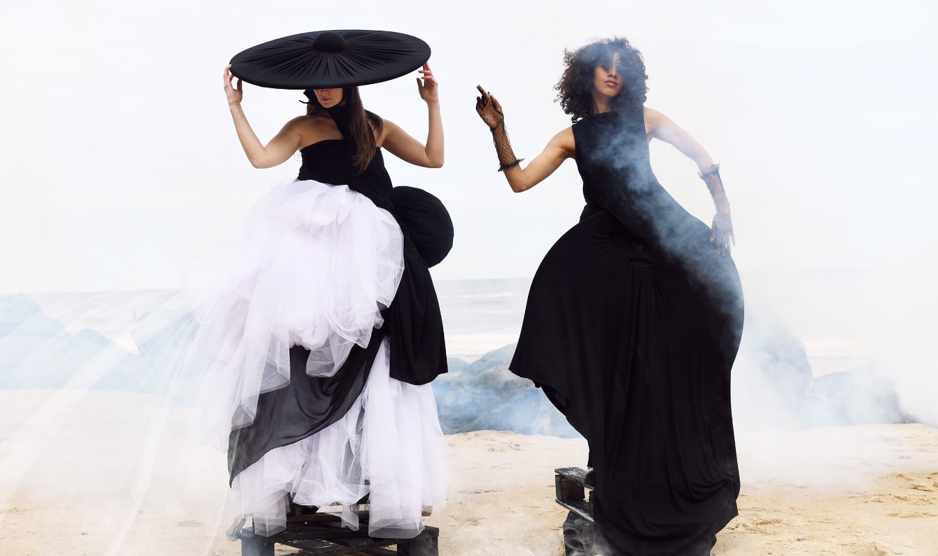 Two women in avant-garde dresses and headpieces pose on a foggy beach, collaborating with a fashion brand. One dress is black and white, the other black. Both women appear to be standing on platforms.