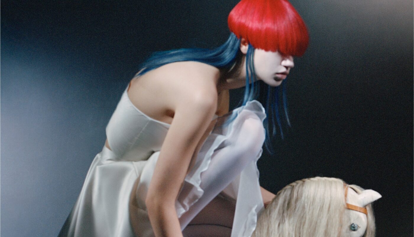 A woman with red hair over her face and blue hair underneath crouches in a white dress next to a toy rocking horse, showcasing a unique blend of colour and style that epitomises avant-garde fashion design.