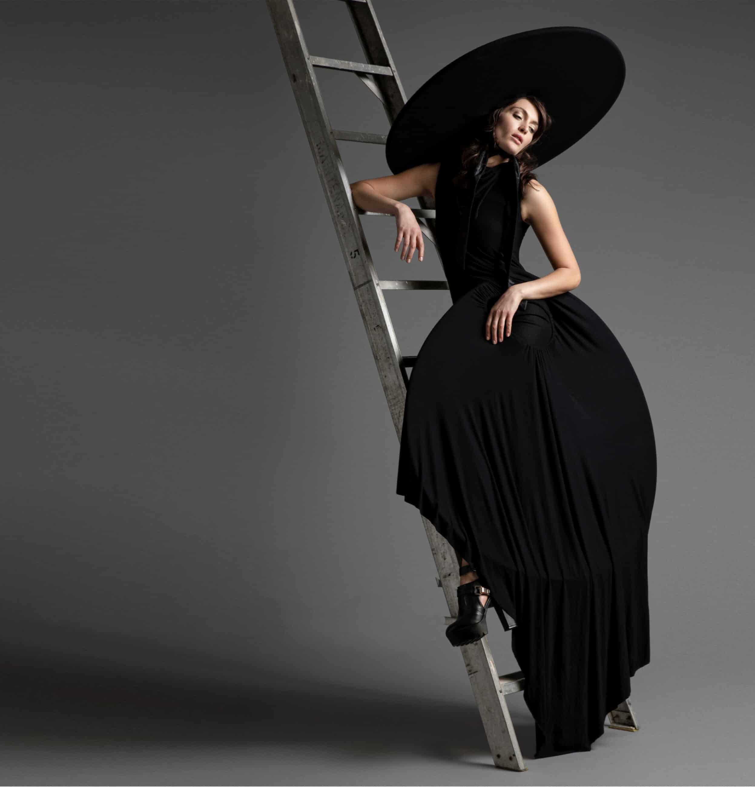 A woman in an elegant black dress and large hat, embodying the sophistication taught at HASOU Fashion Academy, leans on a tall ladder against a grey background.