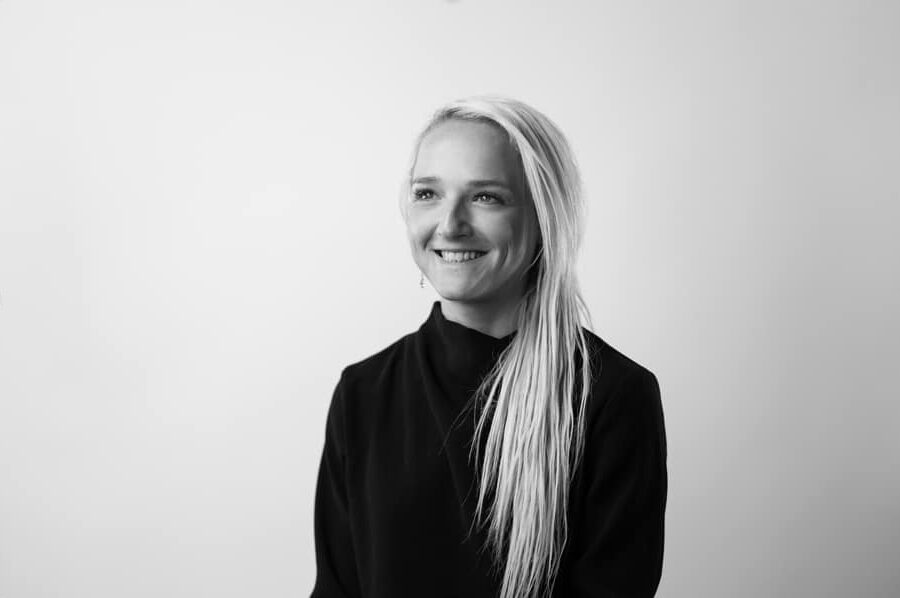 A person with long, light-coloured hair and wearing a dark, high-collared top smiles whilst looking slightly to the side, embodying the chic style often highlighted in online fashion courses. The background is plain and light-coloured.