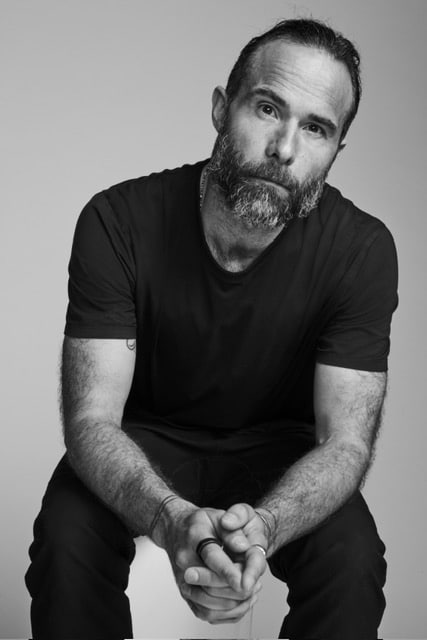 Black and white photo of a man with a beard and short hair, wearing a plain dark T-shirt and dark trousers, sitting and holding his hands together whilst looking at the camera—a perfect look for promoting online fashion courses.