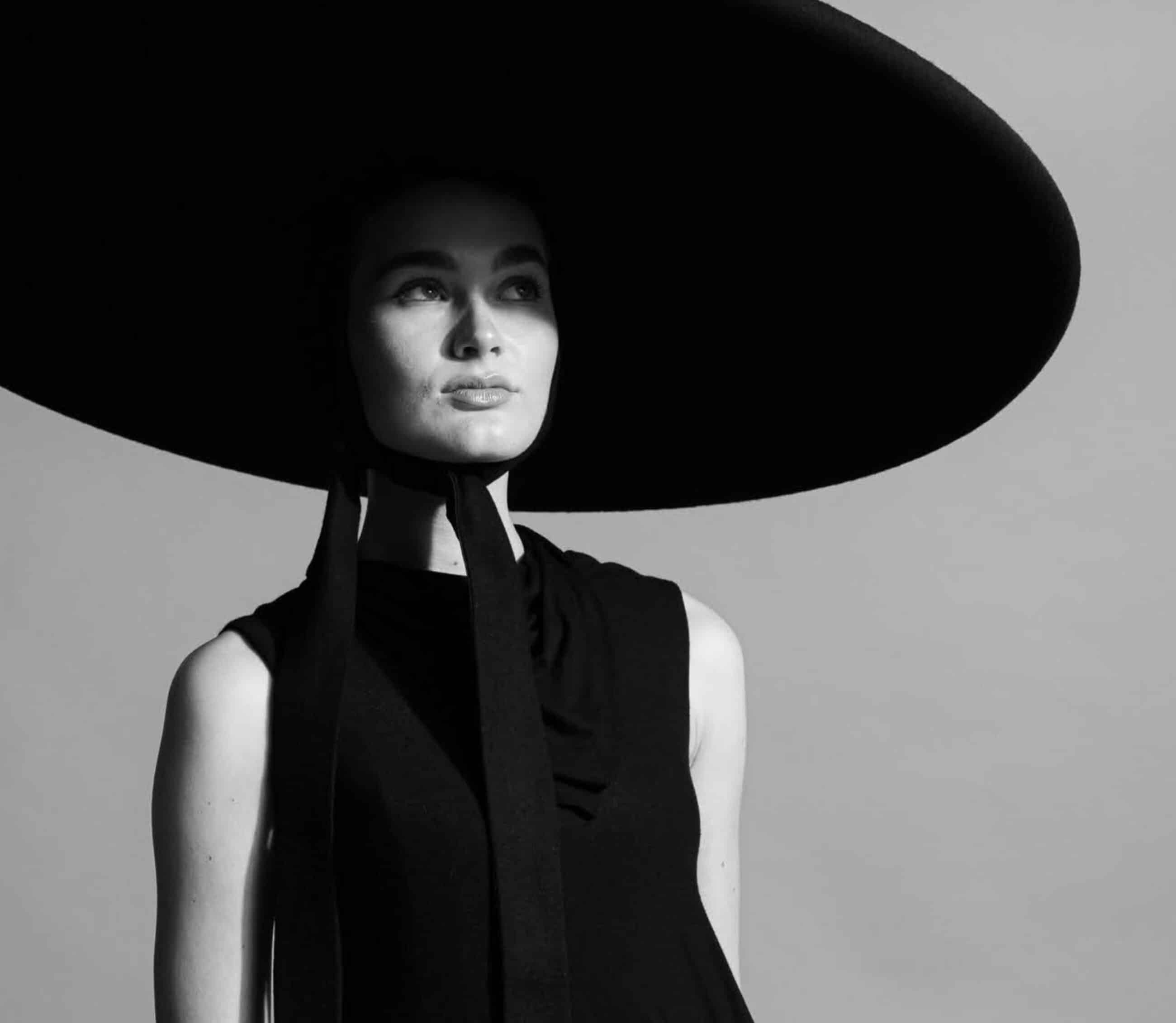 A woman in a sleeveless black dress wears a large, wide-brimmed black hat tied under her chin. She looks slightly upwards against a plain background. The photo, reminiscent of stylish elegance taught in online fashion courses, is in black and white.