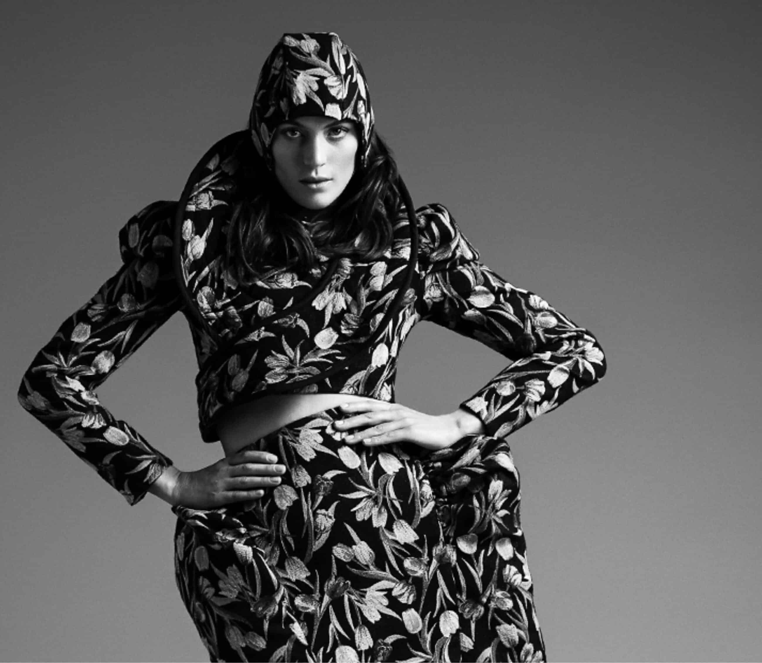 A person wearing an intricately patterned outfit poses with hands on hips, showcasing a matching hat, jacket, and skirt. The photo is in black and white, offering help choosing the perfect ensemble.