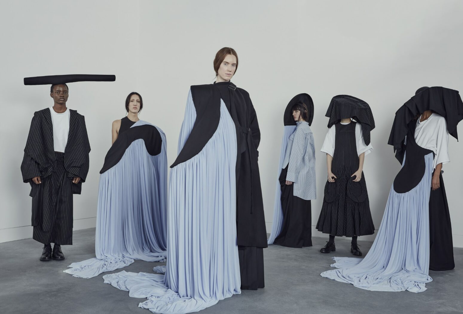 Six individuals are standing, each wearing elaborate, modern black and pale blue outfits with flowing fabric and sculptural elements against a plain background, embodying the essence of careers preparation through their distinct and sophisticated attire.