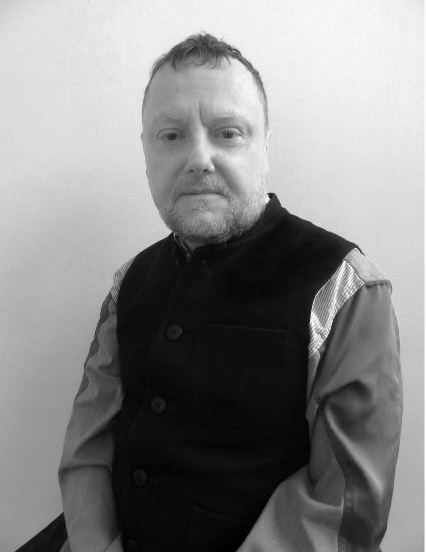 A black-and-white portrait of a man with short hair and a beard, wearing a buttoned shirt and waistcoat, seated and facing the camera against a plain background.