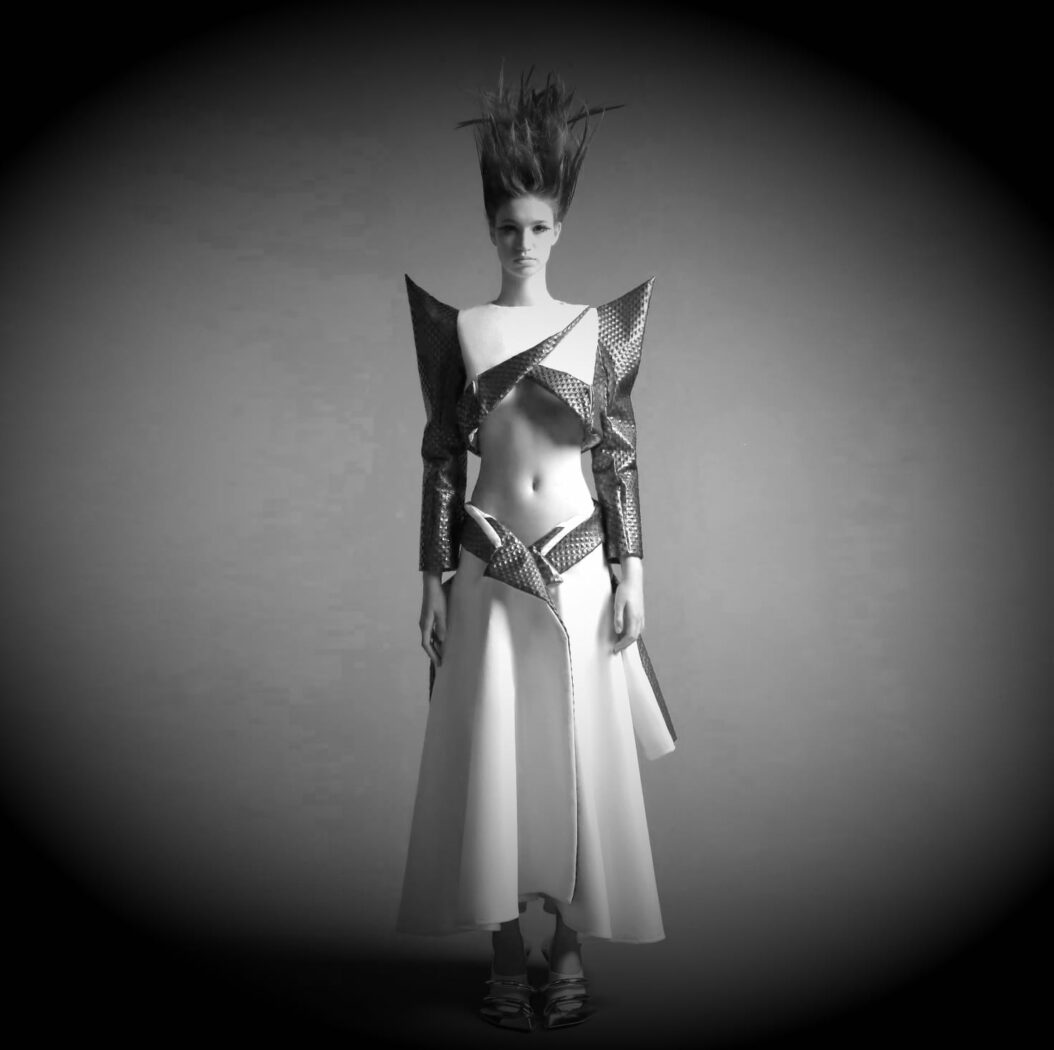 A black-and-white image of a model in a futuristic outfit features sharp, angular shoulder pieces, a cropped top exposing the midriff, and a long, structured skirt. The model's hair is styled high and dramatically, adding to the avant-garde appearance—perfect for those inspired by online fashion courses.