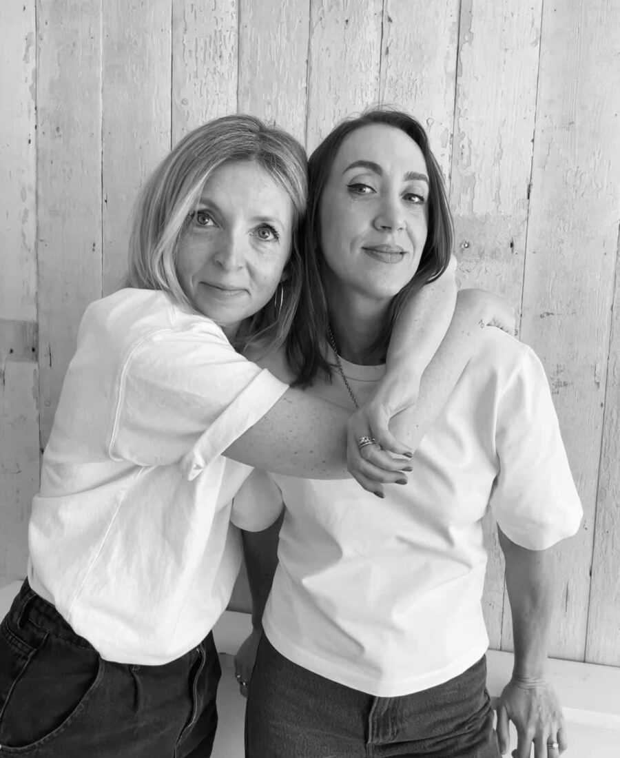 Two women in white shirts stand side by side against a wooden wall, with the woman on the left hugging the other. Both are smiling subtly. Black and white photo. This timeless image could easily grace the cover of any online fashion course, exuding elegance and simplicity.