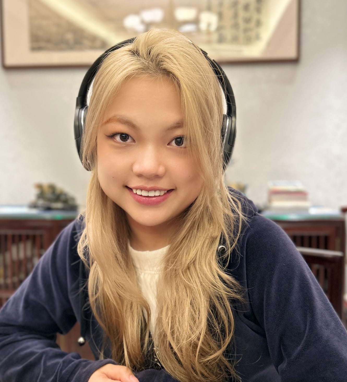 A person with long blonde hair is smiling and wearing headphones, likely engaged in one of their online fashion courses. They are indoors, with a shelf and a framed picture in the background.