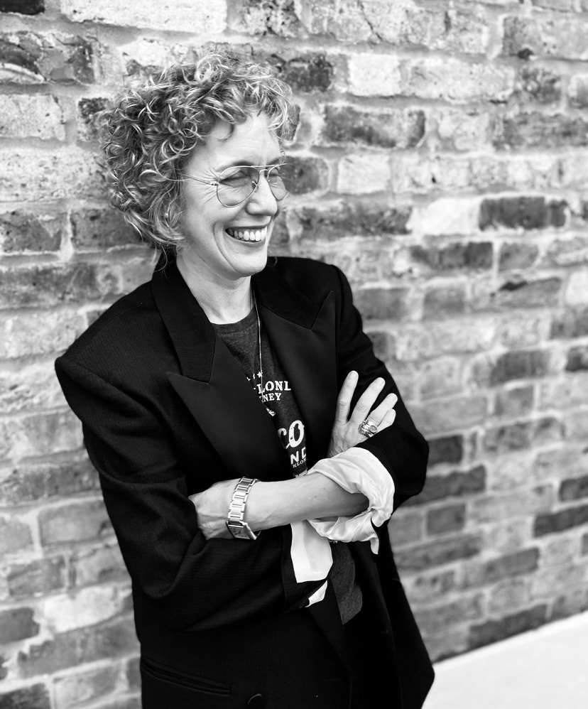 A person with short, curly hair and glasses stands against a brick wall, smiling with arms crossed, dressed in a blazer and casual clothes, embodying the style taught in online fashion courses.