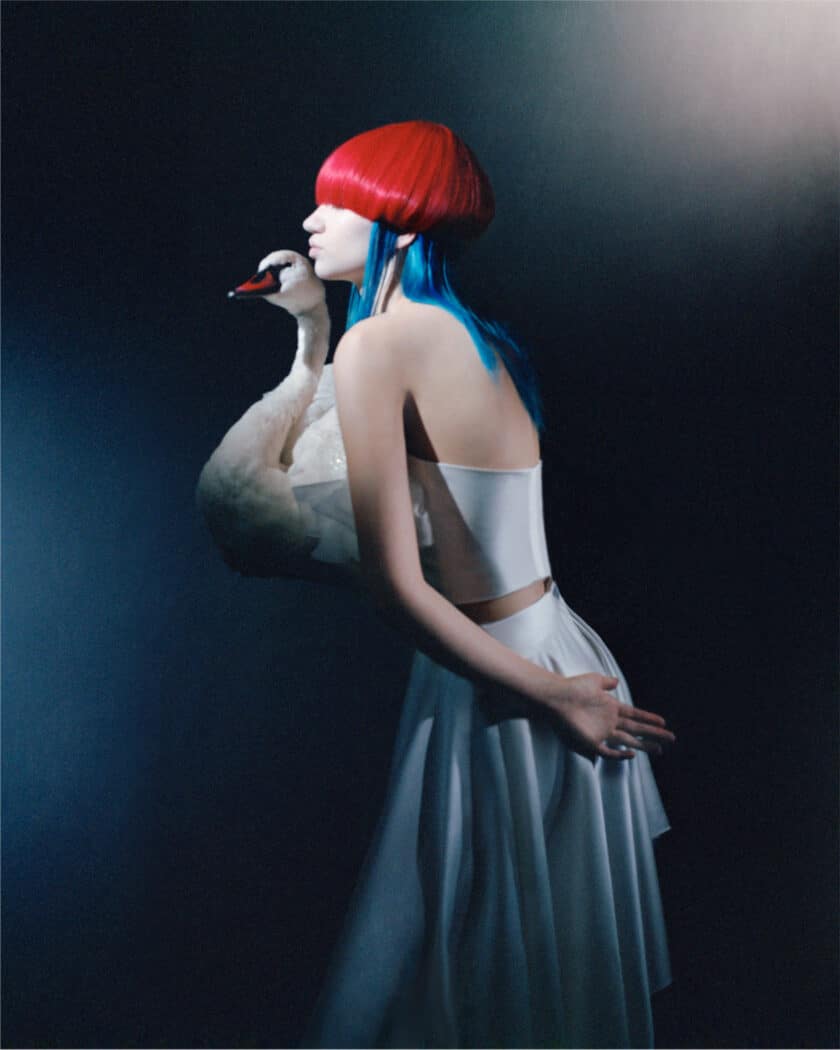 A person with bright red and blue hair, wearing a white outfit, holds a swan against a dark, gradient background, showcasing impeccable fashion design.