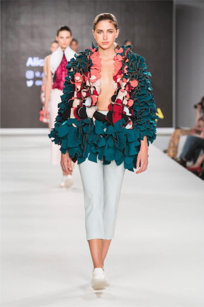 Model walking down the runway, capturing the essence of work in the fashion industry, wearing a unique, textured teal and red jacket with white cropped trousers and white shoes during a fashion show.