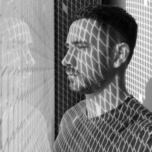 A man stands beside a window with a grid pattern casting shadows on his face and shirt. With a contemplative expression, he gazes out, perhaps reflecting on new trends from his recent online fashion courses.