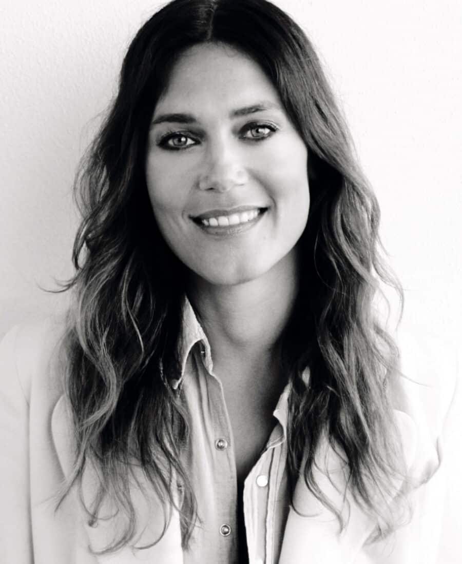 Black and white photo of a woman with long wavy hair, smiling. She is wearing a button-up shirt under a blazer, facing the camera. Her confident look embodies the essence of Online Fashion Courses, combining classic style with modern learning.