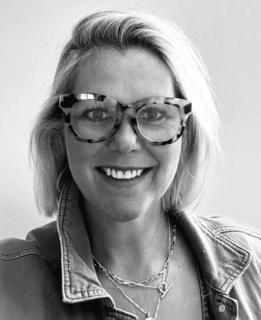 A person with short, light-coloured hair is smiling. They are wearing glasses with a patterned frame and a denim jacket.