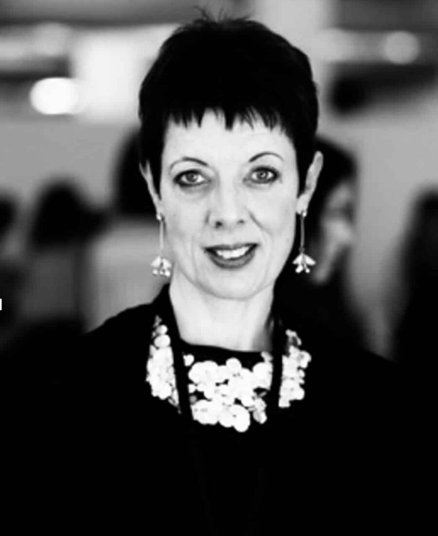 Black and white portrait of a woman with short hair, wearing a necklace and earrings. She smiles slightly with a blurred background of people, reflecting the elegance one might master in online fashion courses.