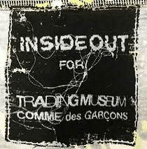 A black and white label displaying the text "INSIDE OUT for TRADING MUSEUM COMME des GARÇONS." The label has a distressed and worn appearance with frayed edges and a visibly textured background.