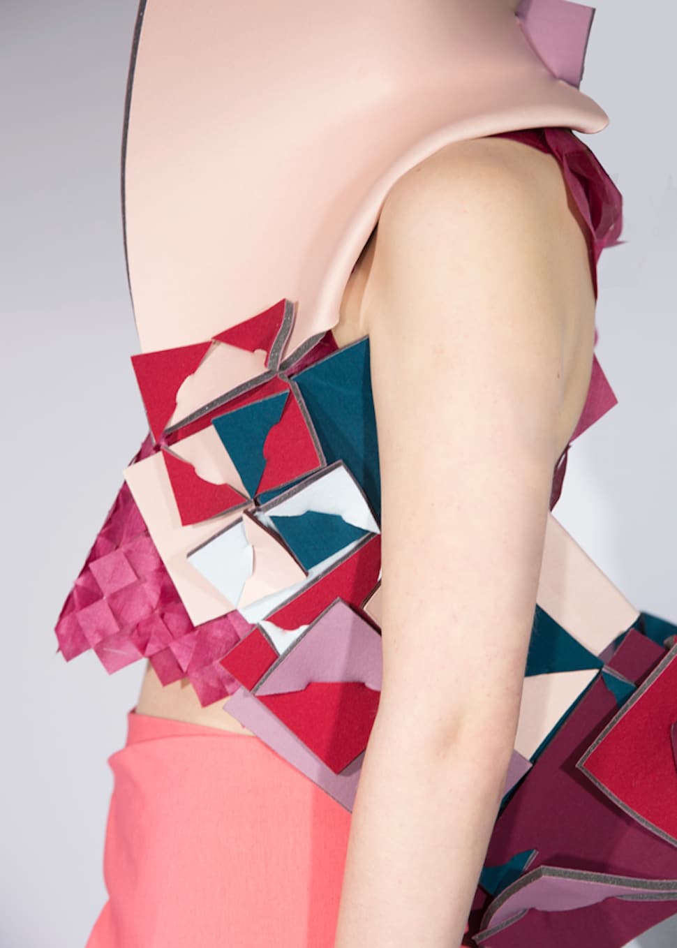 A side view of a person wearing a geometric, multi-coloured garment with pink, red, and teal shapes. The arm and part of the torso are visible against a plain background.