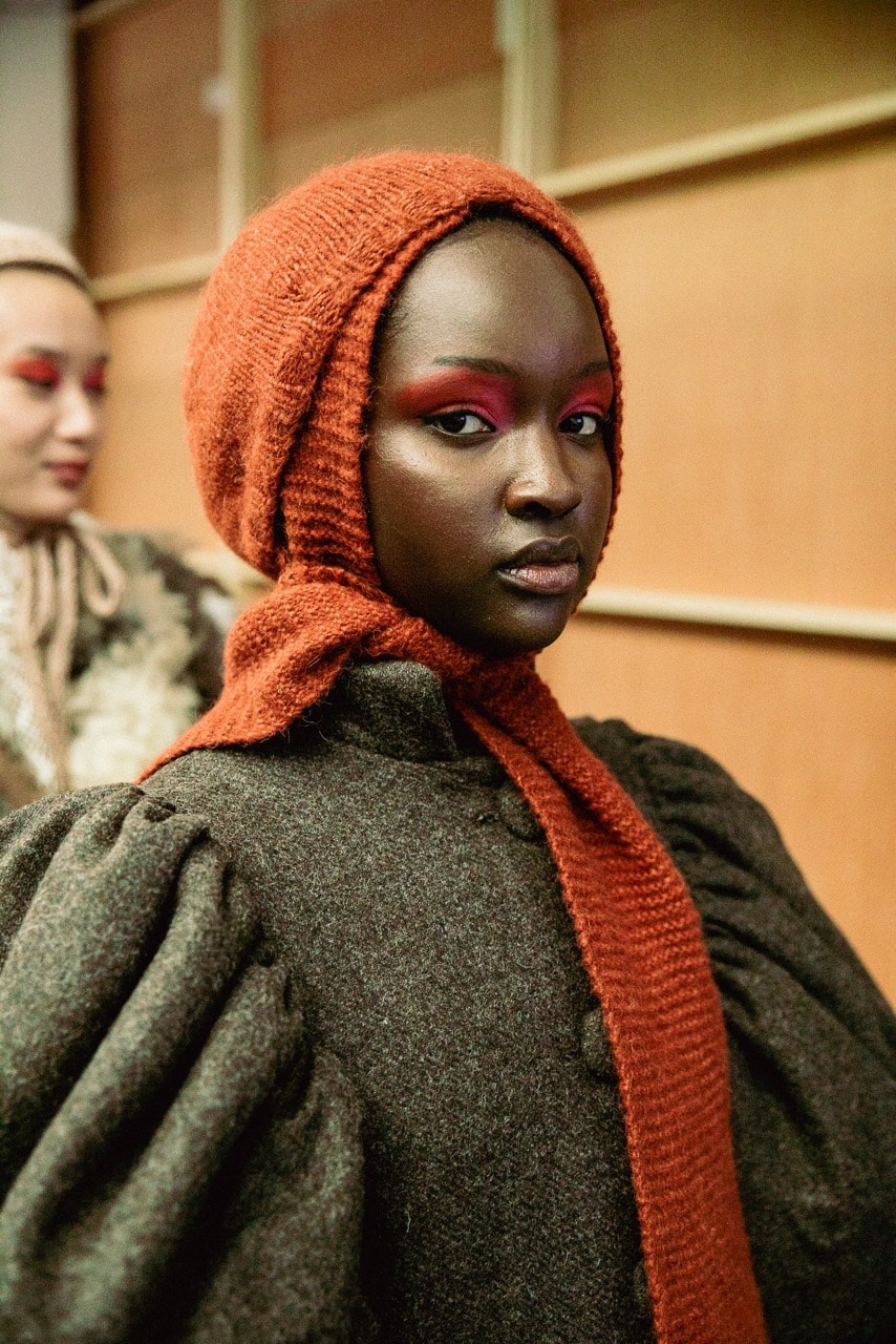 A person wearing a knitted orange headscarf and a dark textured garment looks into the camera, whilst another person in similar clothing is blurred in the background.