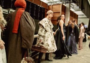 Models backstage in various elaborate outfits, including a floral-patterned cloak and a dark blue dress, preparing for Graduate Fashion Week 2024.