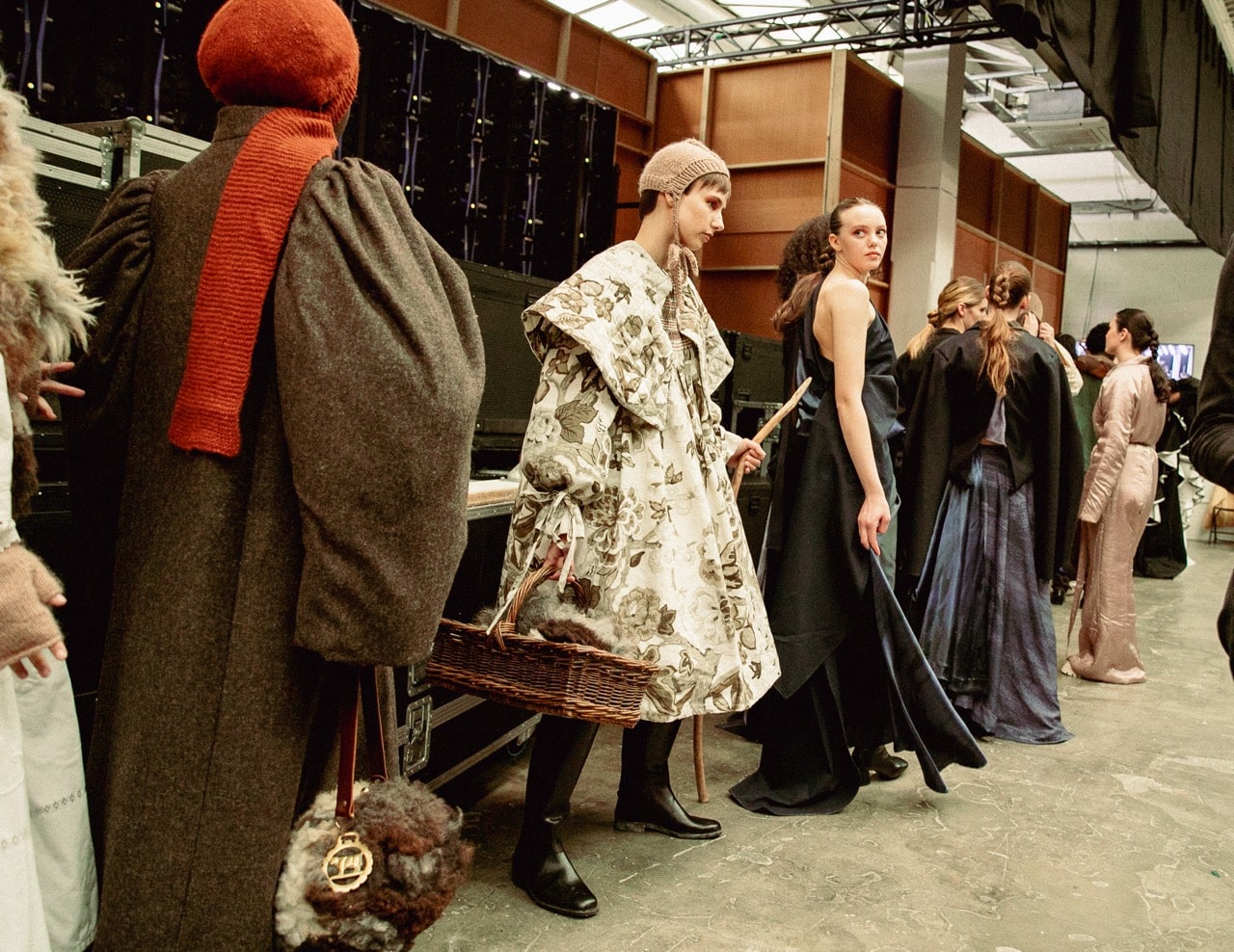 Models backstage in various elaborate outfits, including a floral-patterned cloak and a dark blue dress, preparing for Graduate Fashion Week 2024.