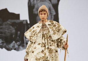 A model strides down the catwalk in a floral-patterned outfit complemented by a voluminous cape. She holds a wooden stick in one hand and carries a wicker basket in the other. Her look is completed with a knitted beanie perched on her head and sturdy black boots on her feet.