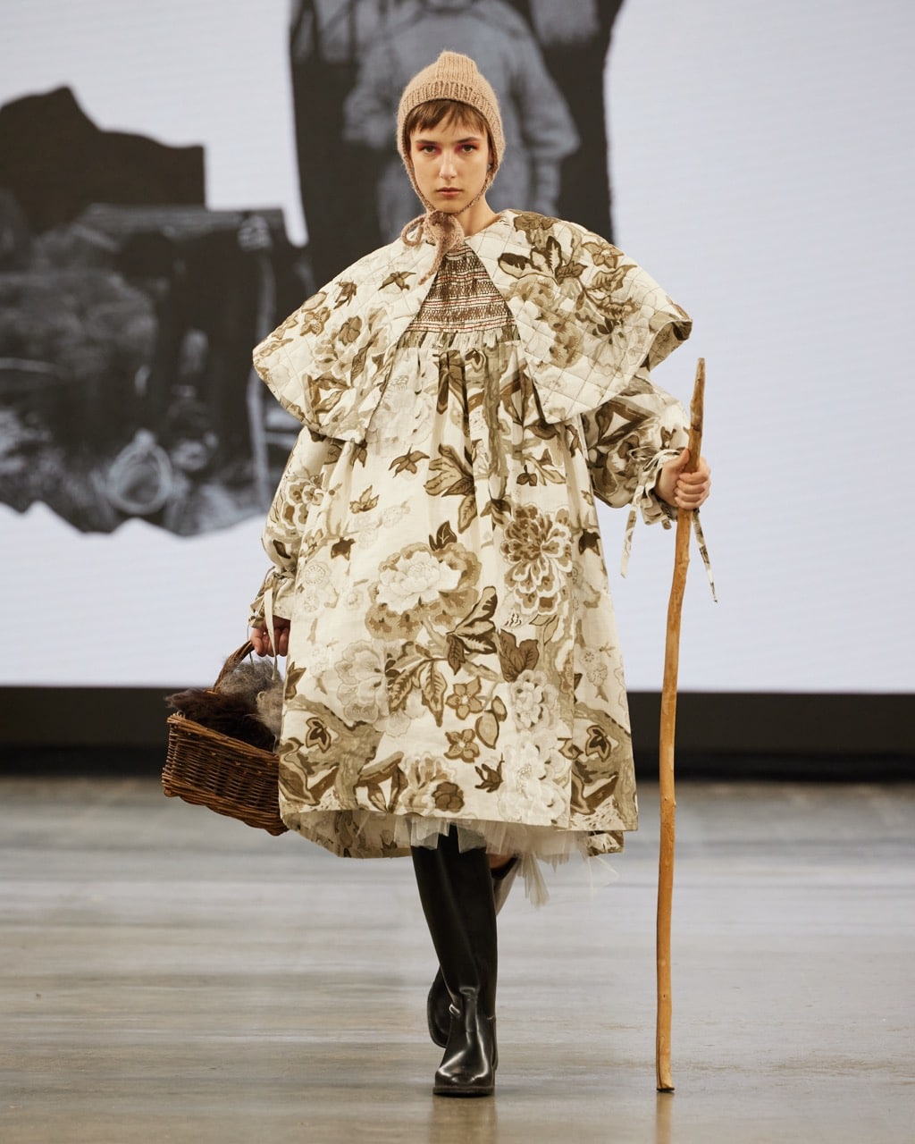 A model strides down the catwalk in a floral-patterned outfit complemented by a voluminous cape. She holds a wooden stick in one hand and carries a wicker basket in the other. Her look is completed with a knitted beanie perched on her head and sturdy black boots on her feet.