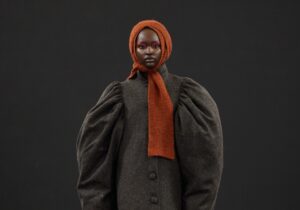 Person wearing a dark heavy coat with voluminous sleeves and an orange headscarf, carrying a furry handbag, walking on a runway with a dark background during Graduate Fashion Week 2024.