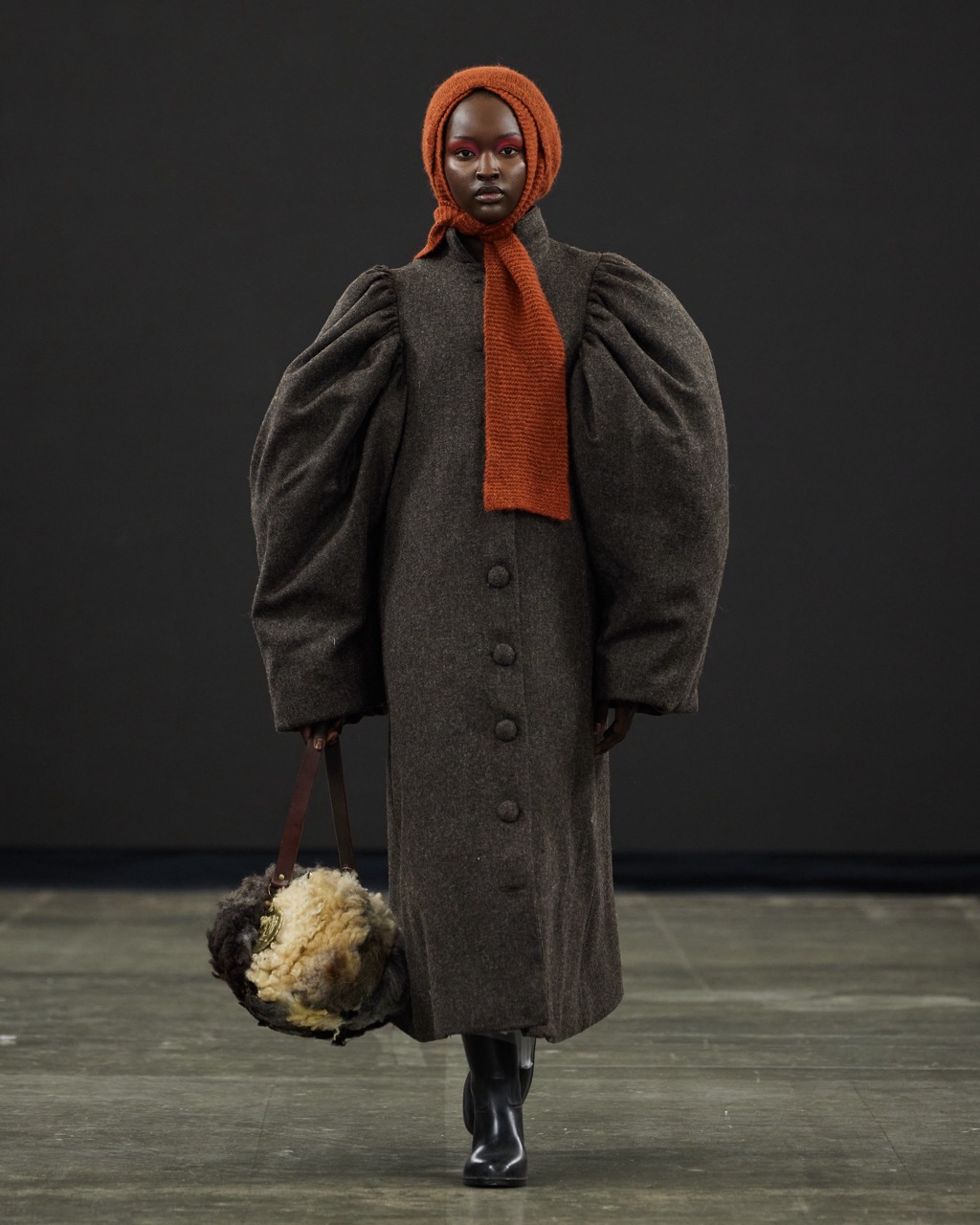 Person wearing a dark heavy coat with voluminous sleeves and an orange headscarf, carrying a furry handbag, walking on a runway with a dark background during Graduate Fashion Week 2024.