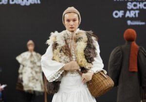 A model walks the catwalk in a white dress, fur waistcoat, and knitted head covering, carrying a wicker basket. Another model is visible in the background.