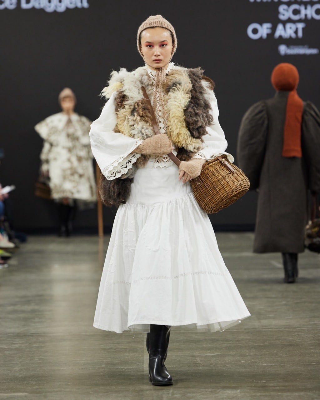 A model walks the catwalk in a white dress, fur waistcoat, and knitted head covering, carrying a wicker basket. Another model is visible in the background.