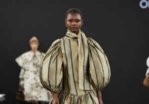 A model walks the runway wearing a tan and black striped dress with large, puffy sleeves and a full, gathered skirt. She is also wearing black boots.