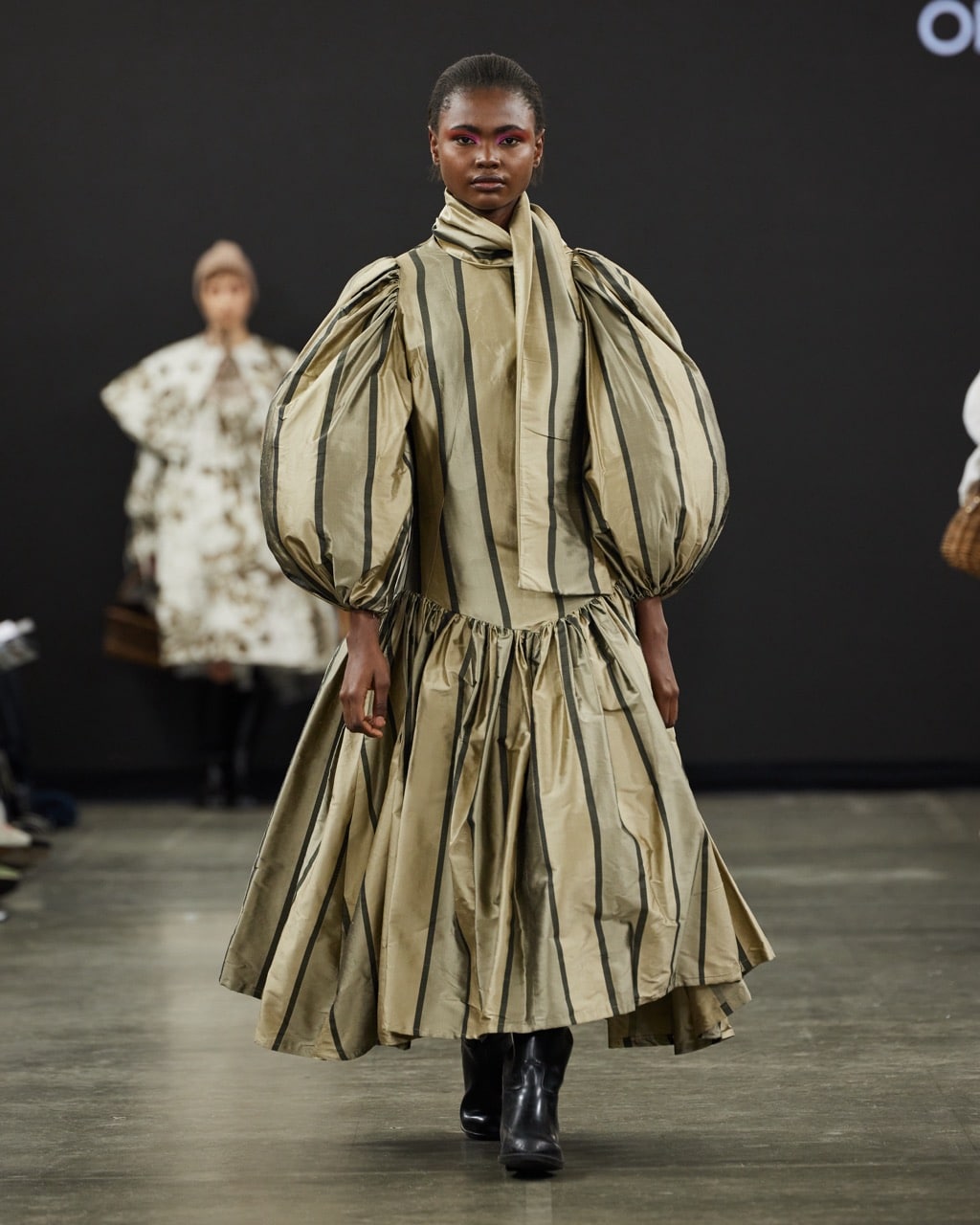 A model walks the runway wearing a tan and black striped dress with large, puffy sleeves and a full, gathered skirt. She is also wearing black boots.