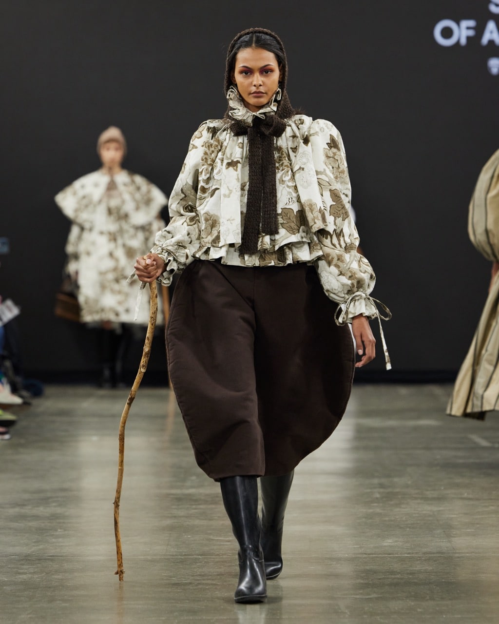 A model on a runway wears a patterned blouse, a decorative scarf, loose brown trousers, and black knee-high boots, holding a wooden stick. Another model in a similar outfit is in the background.