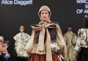 Model walks runway in Graduate Fashion Week 2024 wearing a beige headwrap, a pleated top with puffy sleeves, a large bow, and a red skirt with a brown abstract design. Audience and other models are visible in the background.