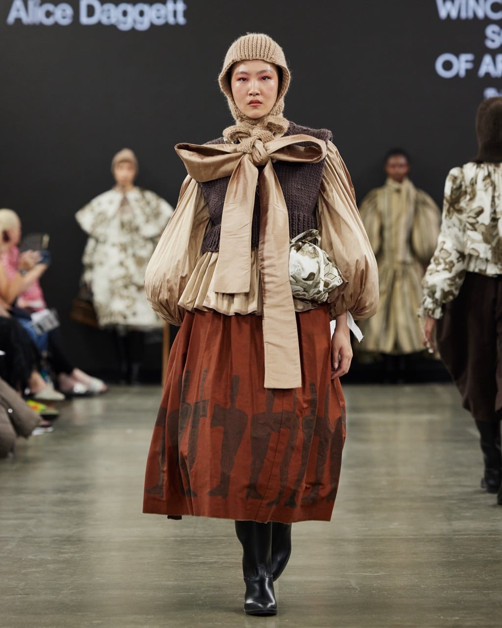 Model walks runway in Graduate Fashion Week 2024 wearing a beige headwrap, a pleated top with puffy sleeves, a large bow, and a red skirt with a brown abstract design. Audience and other models are visible in the background.