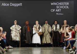 Six models display fashion designs on a runway at Graduate Fashion Week 2024. Designer Alice Daggett's name is displayed on the backdrop. Audience members are seated on both sides of the runway.