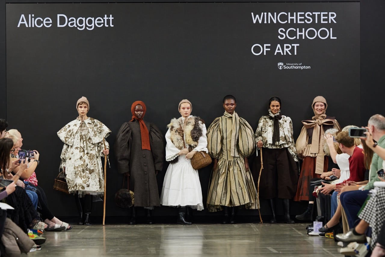 Six models display fashion designs on a runway at Graduate Fashion Week 2024. Designer Alice Daggett's name is displayed on the backdrop. Audience members are seated on both sides of the runway.