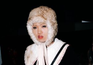 Person wearing a fluffy hat with ear flaps, a white lace collar, a black coat, and matching furry hand accessory, standing against a dark background.