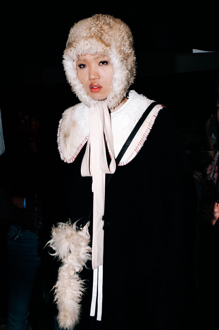 Person wearing a fluffy hat with ear flaps, a white lace collar, a black coat, and matching furry hand accessory, standing against a dark background.