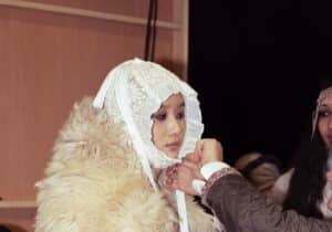 A person wears a detailed outfit with a large fur collar and a lace bonnet whilst another adjusts the bonnet ties.