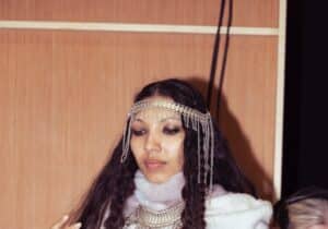 A person with long curly hair is dressed in a layered outfit with a headpiece and necklace. They are posing with one hand raised, standing indoors with a wooden backdrop. Another person is in the background.