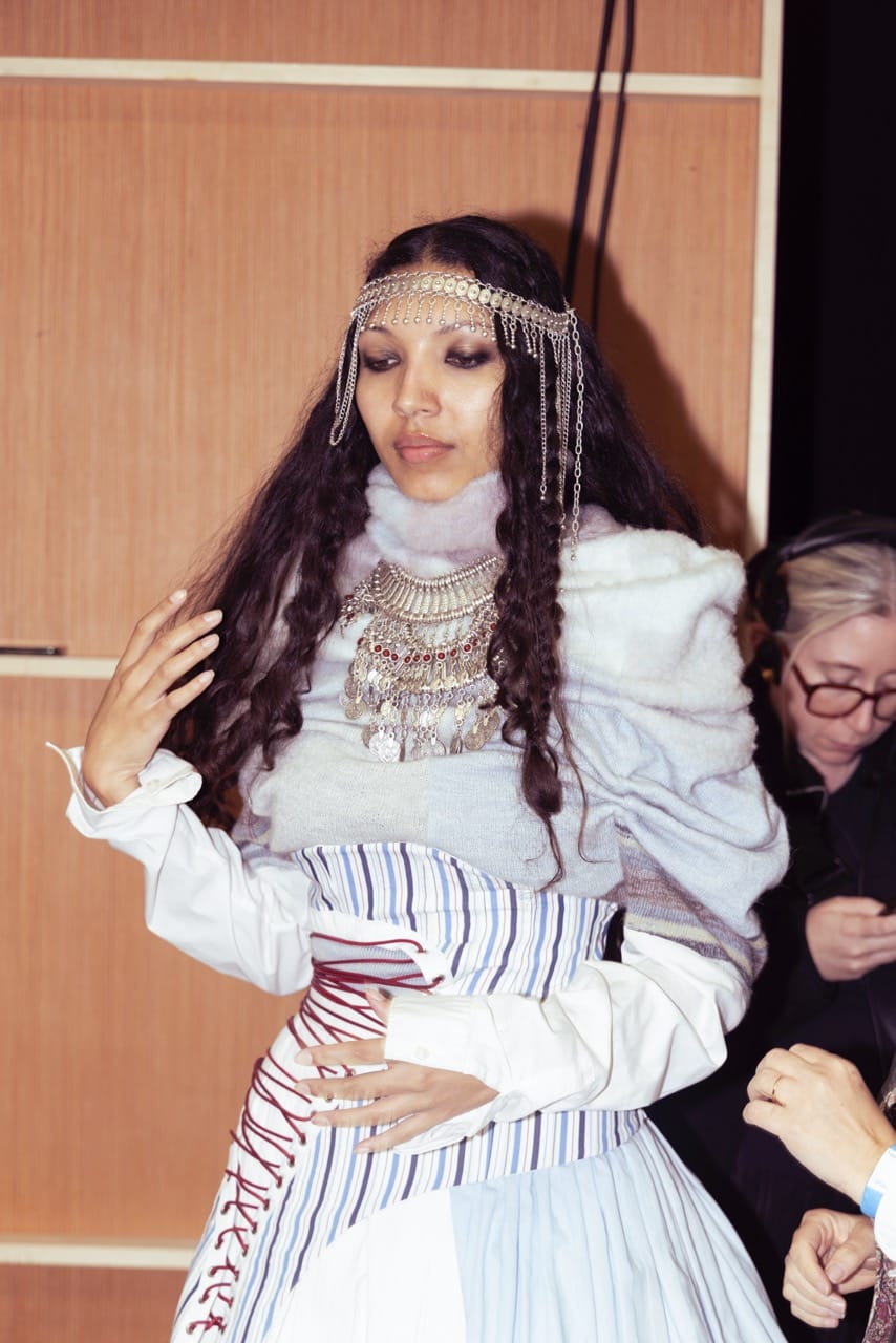 A person with long curly hair is dressed in a layered outfit with a headpiece and necklace. They are posing with one hand raised, standing indoors with a wooden backdrop. Another person is in the background.