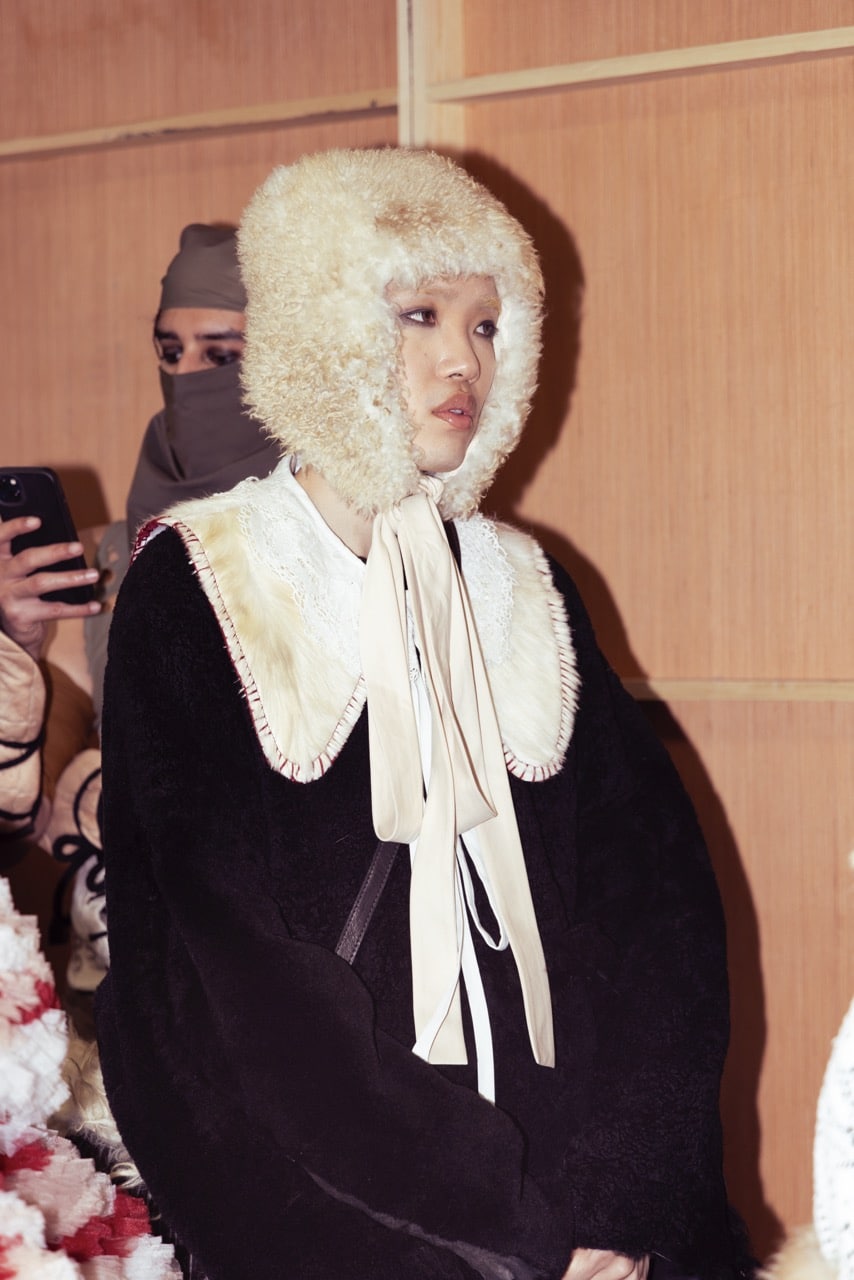 A person wearing a fluffy headpiece, ribbon collar, and dark clothing sits indoors. Another person behind them is holding a smartphone.