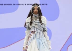 A model walks the runway at Graduate Fashion Week 2024 wearing a layered dress with light blue and white tones, a blue shoulder bag, and a beaded headpiece against a backdrop with abstract designs.