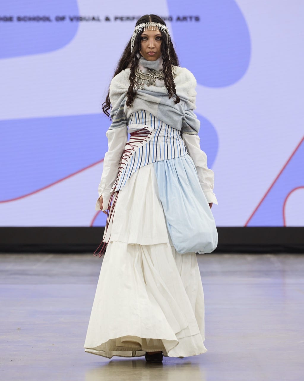 A model walks the runway at Graduate Fashion Week 2024 wearing a layered dress with light blue and white tones, a blue shoulder bag, and a beaded headpiece against a backdrop with abstract designs.