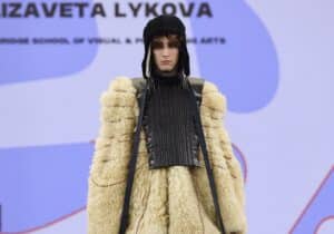 A model walks the runway wearing an eclectic outfit comprised of a shaggy fur coat, a layered skirt, and a black armoured vest, with a knit headpiece.