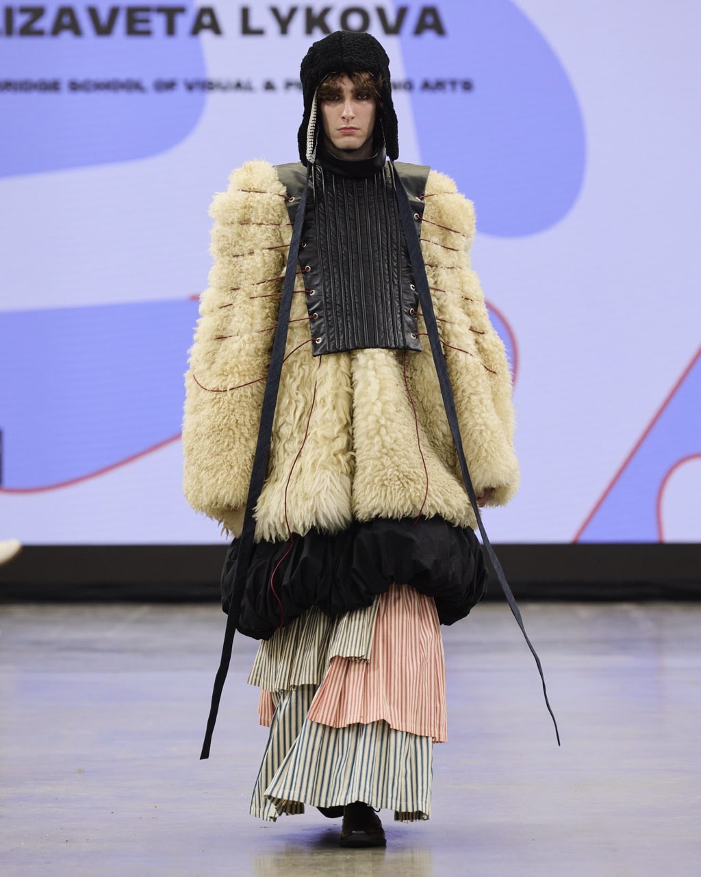 A model walks the runway wearing an eclectic outfit comprised of a shaggy fur coat, a layered skirt, and a black armoured vest, with a knit headpiece.