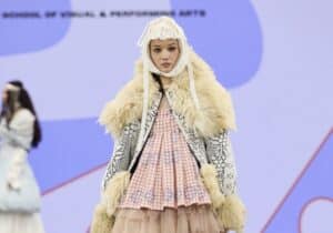 A model walks the runway in a layered outfit with a fur-trimmed jacket, a pink-chequered top, and patterned white and beige skirts during Graduate Fashion Week 2024.